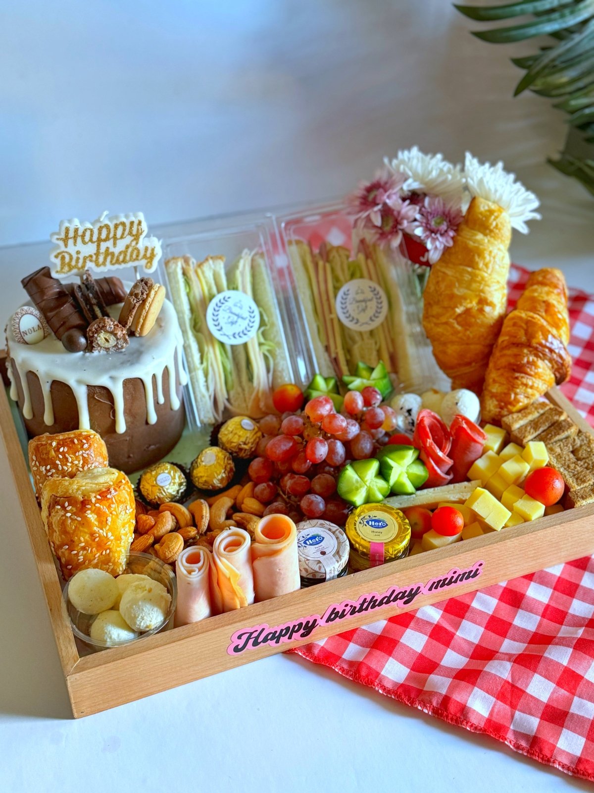A close-up of the birthday breakfast gift, showcasing a delicious chocolate cake topped with Kinder Bueno and macarons, alongside an assortment of gourmet cheeses, fresh grapes, and premium nuts.