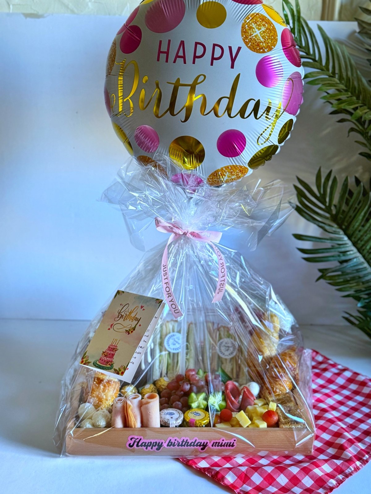 A beautifully arranged birthday breakfast tray featuring a Nola Kinder Bueno Bomb Cake, croissants, cheeses, fresh fruits, nuts, and a Happy Birthday balloon, elegantly wrapped for gifting.