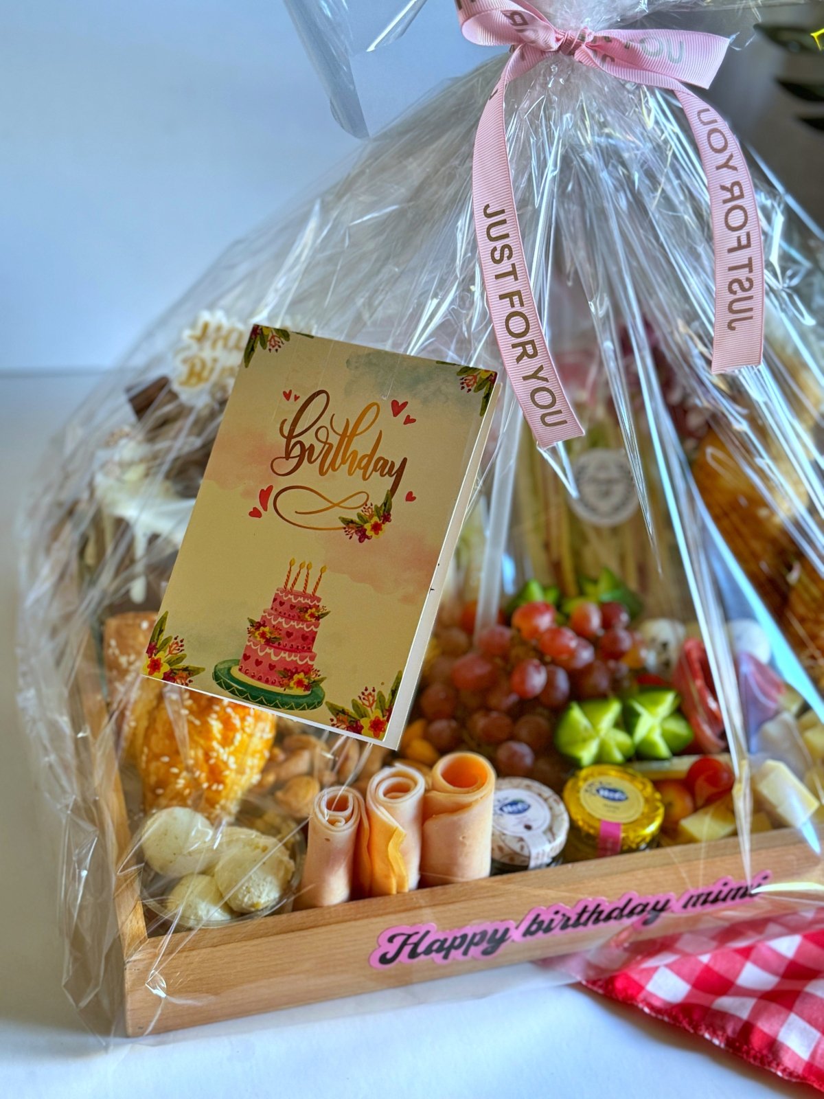 A stylish birthday breakfast tray including club sandwiches, croissants, Ferrero Rocher chocolates, cheese, and a cake, wrapped beautifully with a birthday tag and pink ribbon.