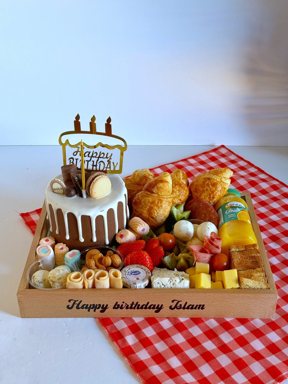 A beautifully arranged wooden birthday tray featuring croissants, cheese cubes, labneh balls, mixed nuts, fresh fruits, and a Nola Kinder Bueno Bomb Cake, placed on a red gingham cloth.