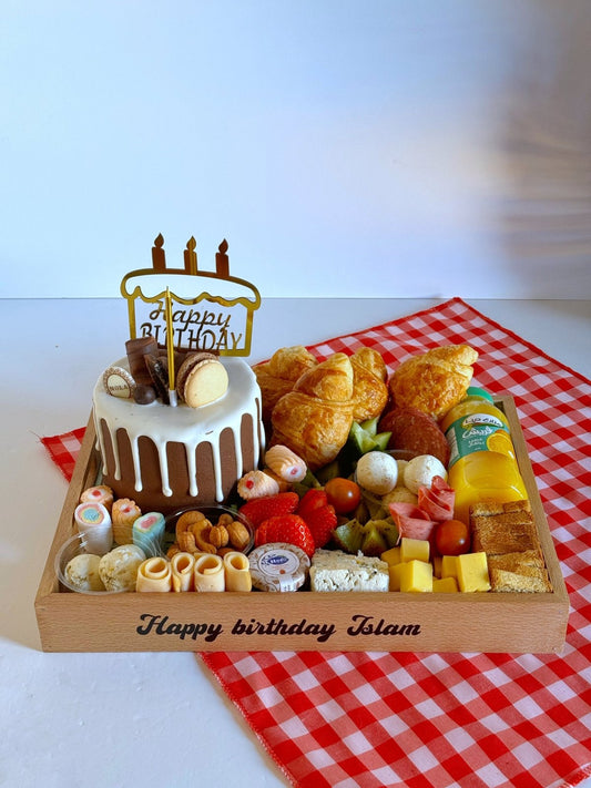 A beautifully arranged wooden birthday tray featuring croissants, cheese cubes, labneh balls, mixed nuts, fresh fruits, and a Nola Kinder Bueno Bomb Cake, placed on a red gingham cloth.