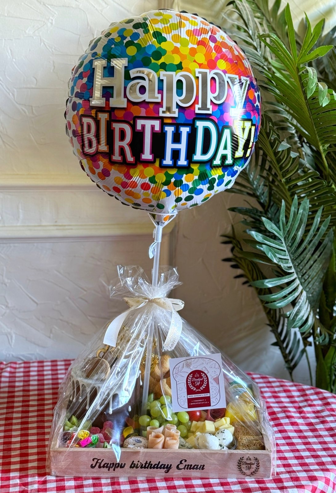 A wrapped birthday gift tray with a colorful "Happy Birthday" balloon, tied with a white ribbon, featuring a selection of gourmet breakfast items.