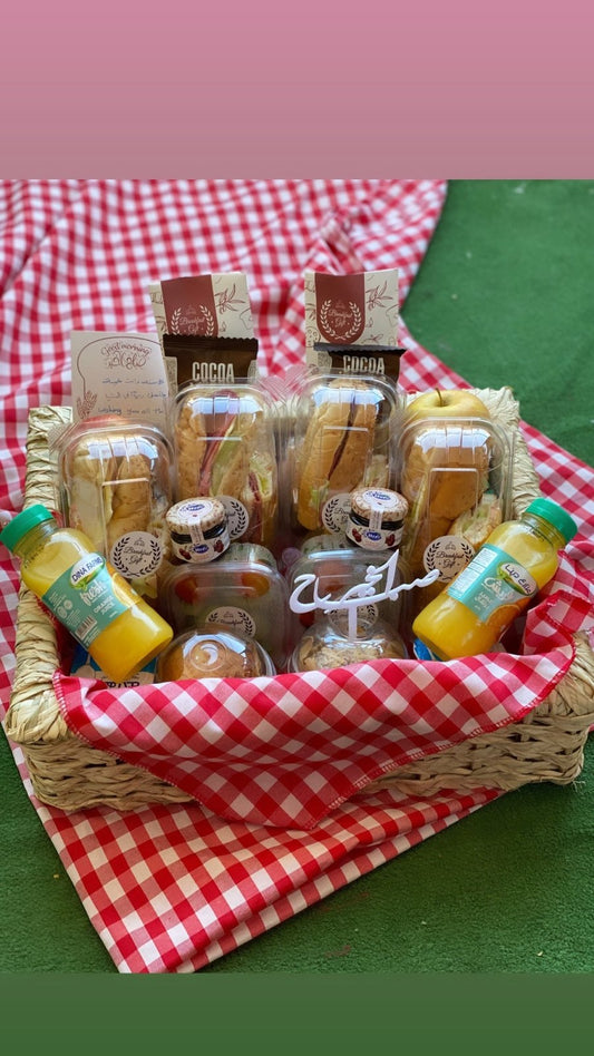 A beautifully arranged wicker basket with a red-and-white checkered cloth, featuring breakfast sandwiches, fresh fruit, juice bottles, and a "صباح الخير" (Good Morning) topper, making it a perfect meal for two.