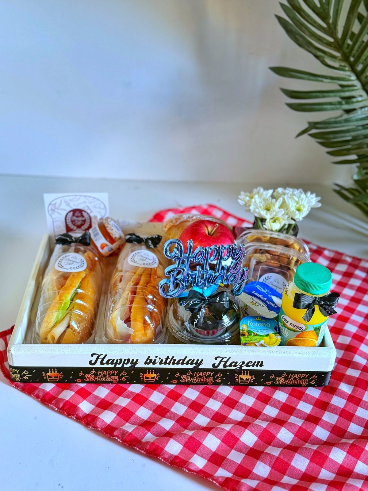 Elegant wooden breakfast tray with sandwiches, toast, fresh fruit, chocolates, juice, and a 'Happy Birthday' topper, accompanied by flowers and a balloon for a delightful gift experience.