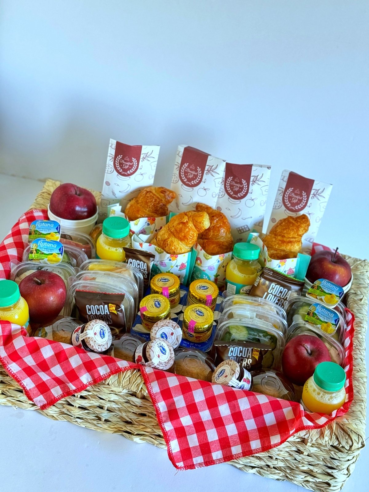 Family Basic Breakfast in a Basket for four – a beautifully arranged wicker tray featuring croissants, roasted toasts, muffin cakes, fresh fruits, tomatoes, cucumbers, natural juices, tea, Hero honey, jam, butter, smoked luncheon, cheddar, and Cocoa Lovers treats. A generous and delicious breakfast set perfect for families or groups to enjoy together.