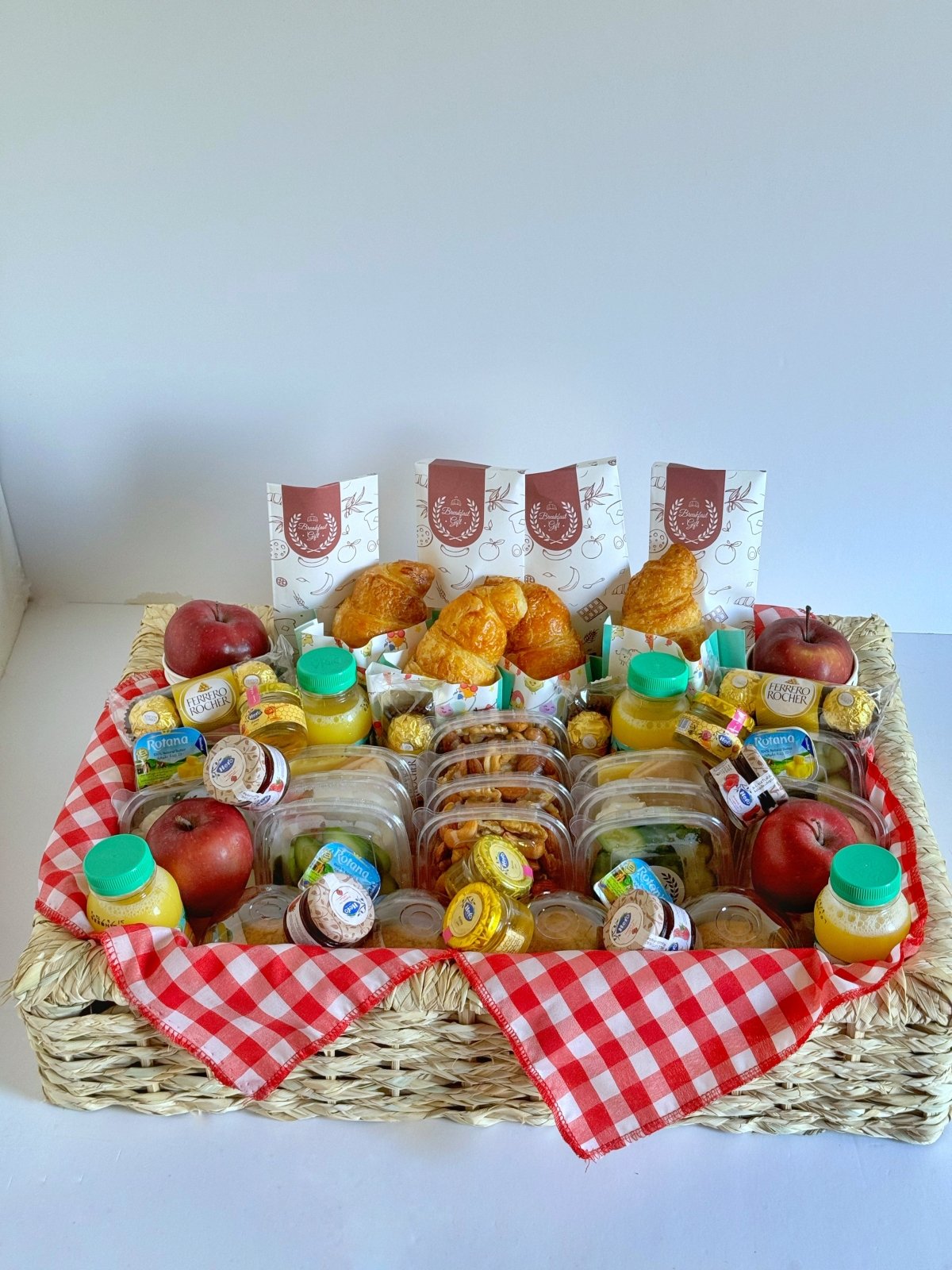Close-up of the Family Breakfast Basket for four, showcasing golden croissants, individual servings of honey, jam, butter, Ferrero Rocher chocolates, fresh fruits, natural juices, and premium cheeses. A luxurious and satisfying breakfast experience in an elegant wicker tray with a checkered mat.