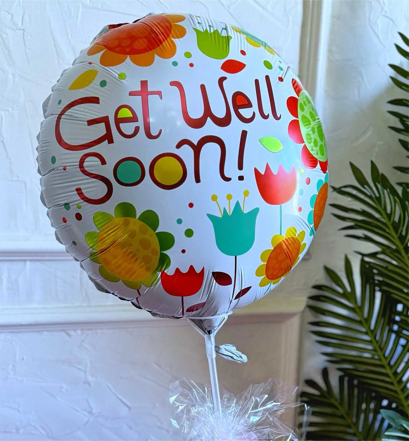 A close-up of a "Get Well Soon" balloon with colorful floral designs, tied to a fruit basket. The vibrant arrangement of fresh fruits and nuts is visible in the background.