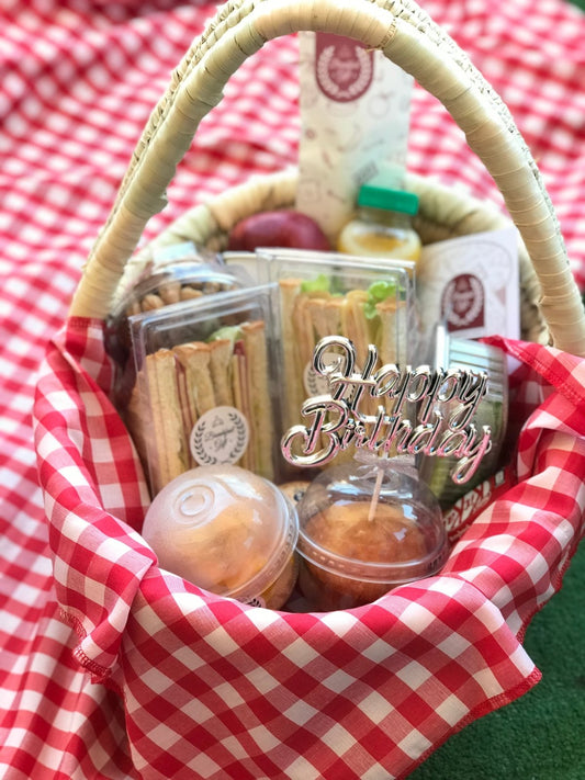A beautifully arranged Happy Birthday Breakfast Basket featuring club sandwiches, a muffin, fresh fruit, a Kit Kat chocolate, and a "Happy Birthday" topper, presented in a wicker basket with a red-and-white checkered cloth.