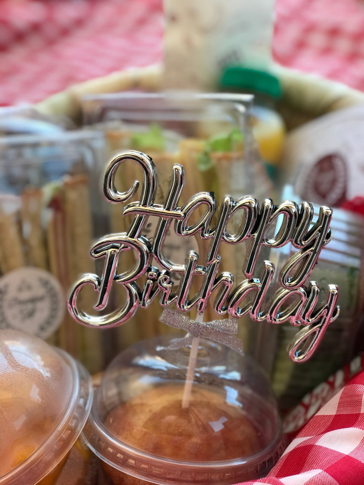 A close-up view of the Happy Birthday Breakfast Basket, highlighting a Happy Birthday topper, sandwich packs, a fruit salad, a muffin, and a tea cup with sugar, all elegantly placed inside a decorative basket.