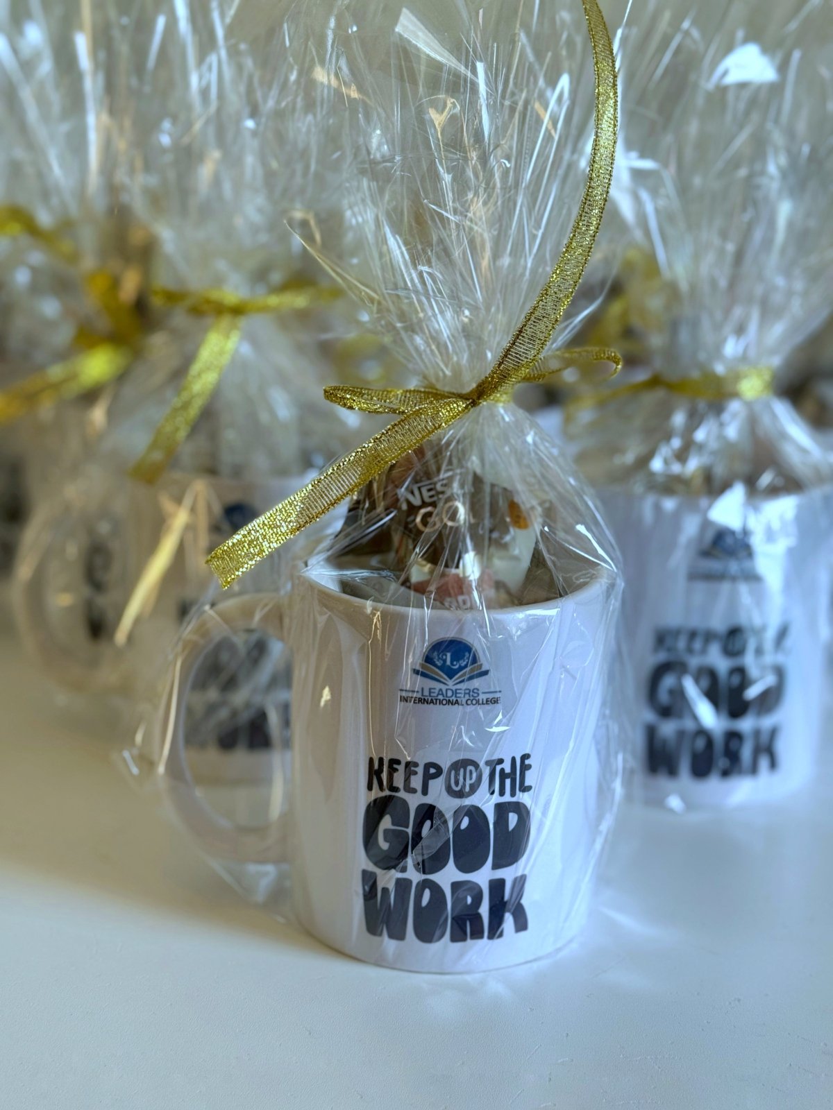 Personalized appreciation mugs wrapped in transparent gift bags with golden ribbons, each featuring a motivational quote and school logo.