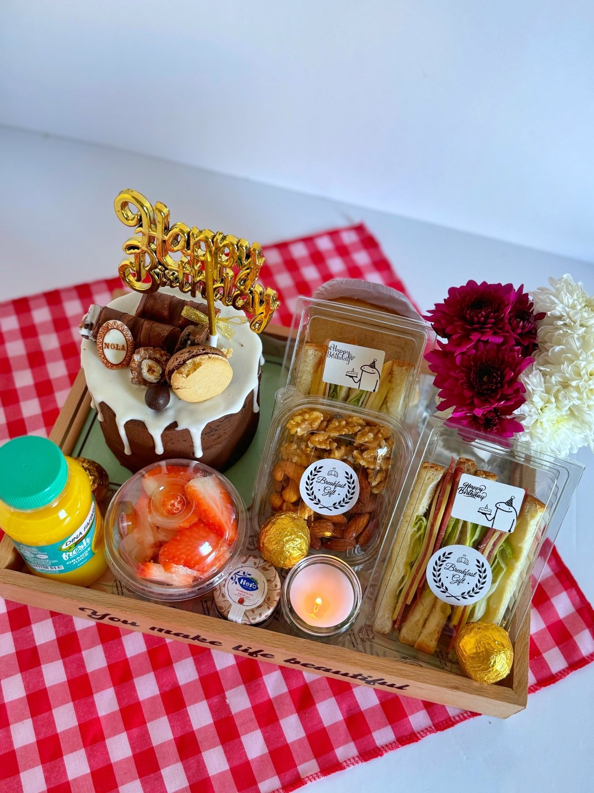 A charming birthday breakfast gift set arranged on a wooden tray, showcasing a Nola chocolate cake, fresh sandwiches, nuts, a candle, and a fruit salad, with vibrant flowers adding an elegant touch.