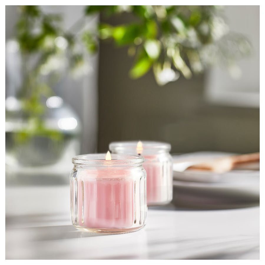 A soft pink scented candle in a clear glass jar, casting a warm and inviting glow—perfect for relaxation, ambiance, or as an elegant addition to a thoughtful gift.