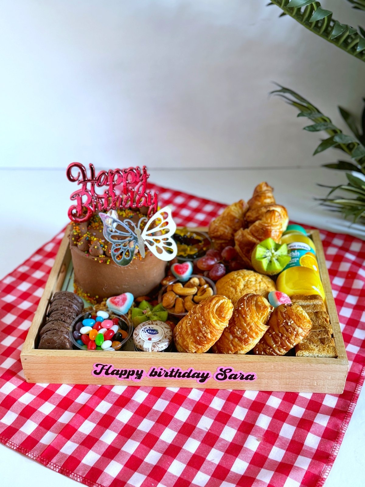 A beautifully arranged Birthday Cake & Bakery gift tray featuring a rich chocolate birthday cake with a red 'Happy Birthday' topper, fresh croissants, pastries, assorted nuts, fruits, and gourmet sweets, served on a charming wooden tray.