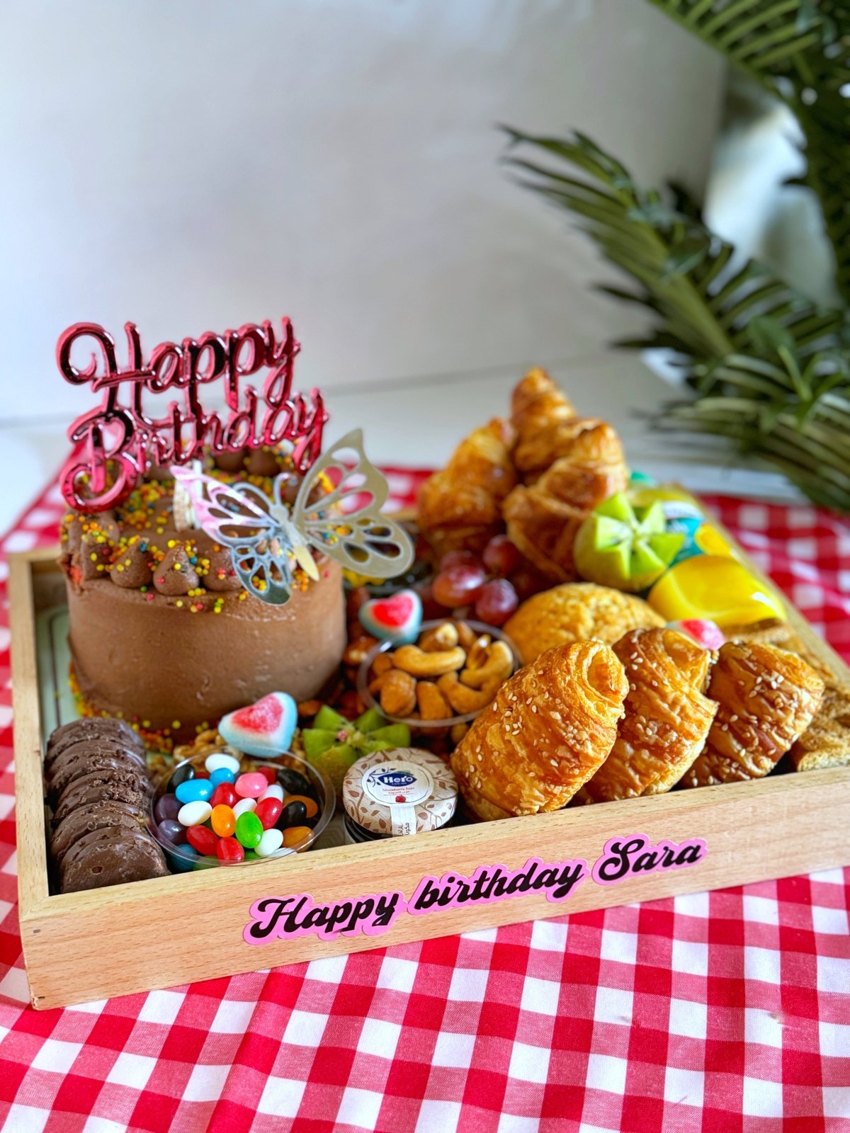 A delightful celebration tray showcasing a chocolate birthday cake, golden croissants, cheese pastries, toasted bread slices, colorful jelly candies, fresh fruits, and a selection of nuts, perfectly curated for a special occasion.
