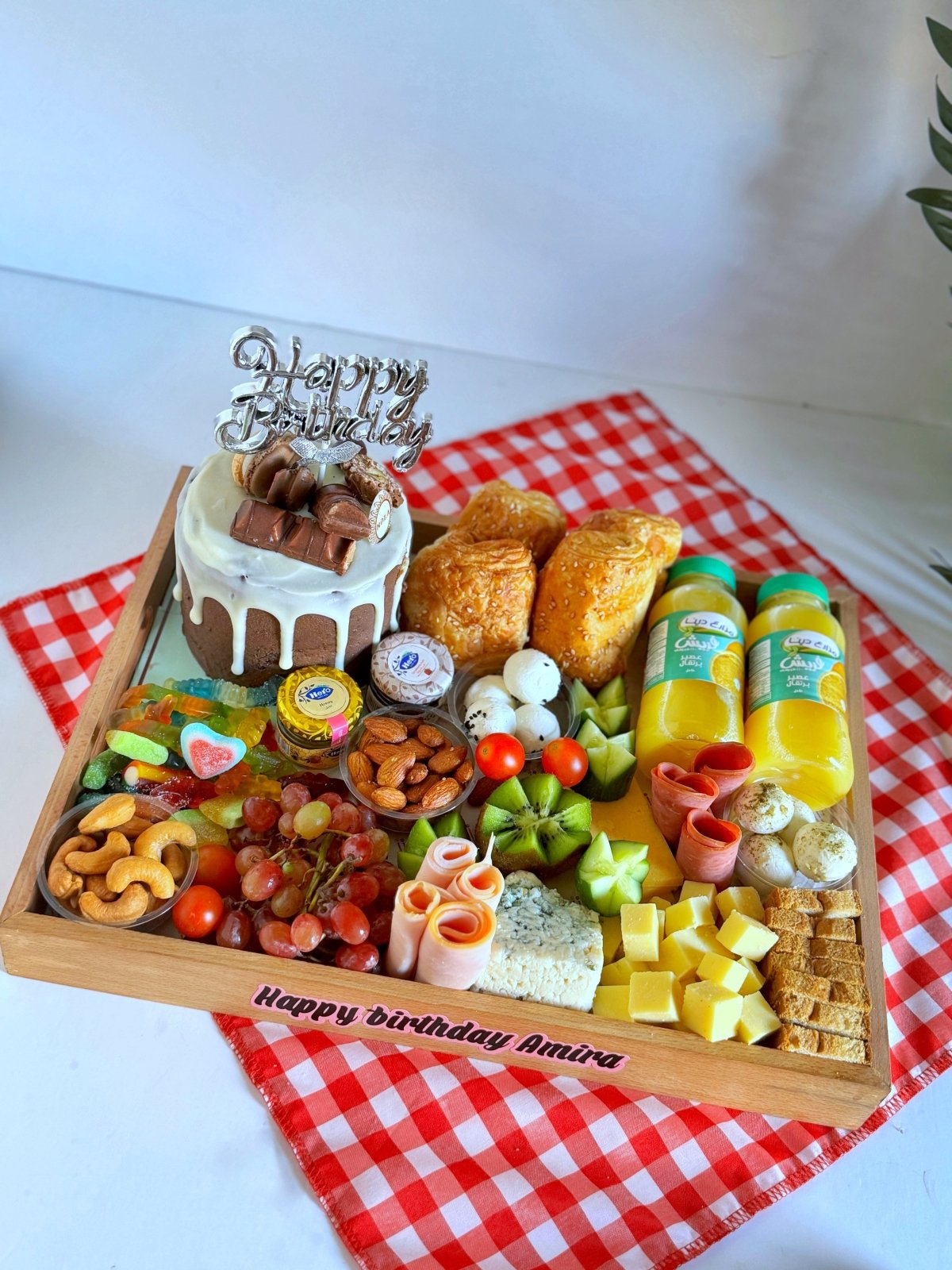 Couples Breakfast Cake & Cheese gift set on a wooden tray featuring a Nola cake with Happy Birthday topper, croissants, toasted bread, cheddar cheese, roquefort cheese, smoked turkey, grapes, kiwi, cucumber, cherry tomatoes, nuts, candy jelly, and two juices, beautifully arranged on a red gingham cloth. Elegantly presented on a wooden tray with a personalized message saying 'Happy Birthday Amira.