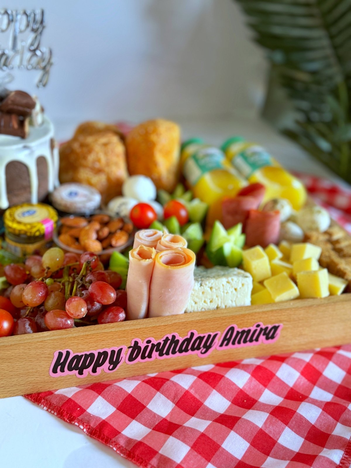 Close-up of the Couples Breakfast Cake & Cheese tray with rolled smoked turkey slices, cheddar cubes, roquefort cheese, grapes, nuts, and candy jelly next to a Nola cake topped with Happy Birthday decor, elegantly presented on a wooden tray with a personalized message saying 'Happy Birthday Amira.