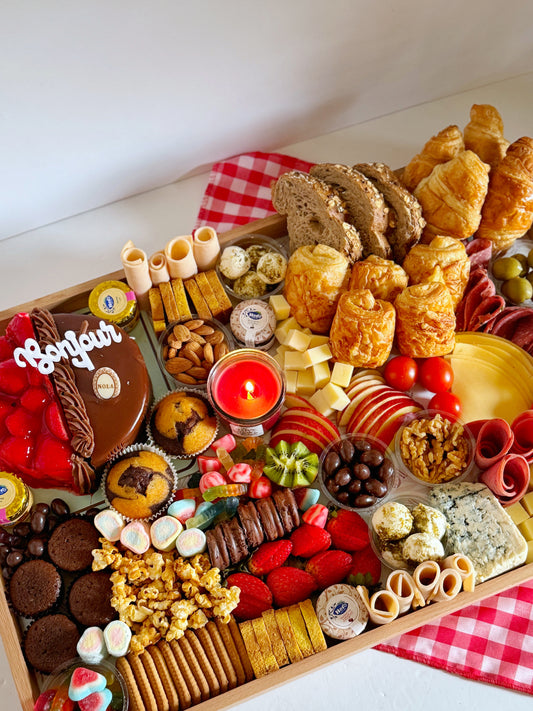 A beautifully arranged wooden tray (60 x 40 cm) featuring an assortment of croissants, whole wheat panini bread, toasted bread, cheddar cheese, blue cheese, turkey salami, roast beef, fresh fruits, nuts, and sweet treats, elegantly displayed on a red checkered cloth.