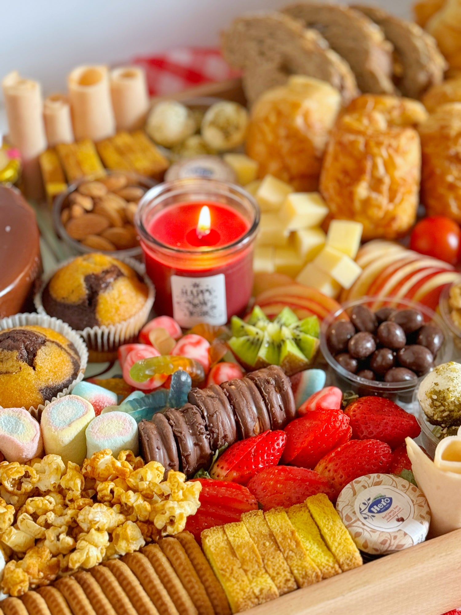 A festive wooden tray featuring caramel popcorn, chocolate-covered peanuts, marshmallows, biscuits, mini cupcakes, muffin cakes, and two jars of jam and honey, perfect for a sweet and savory experience.