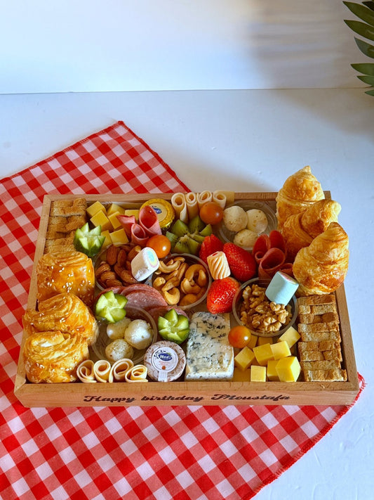 A beautifully arranged wooden breakfast tray featuring an assortment of croissants, cheese-filled puff pastries, toasted bread, cheddar and Gouda cheese cubes, fresh strawberries, grapes, kiwi slices, cherry tomatoes, cucumber slices, and cold cuts including salami, smoked turkey, and roast beef.