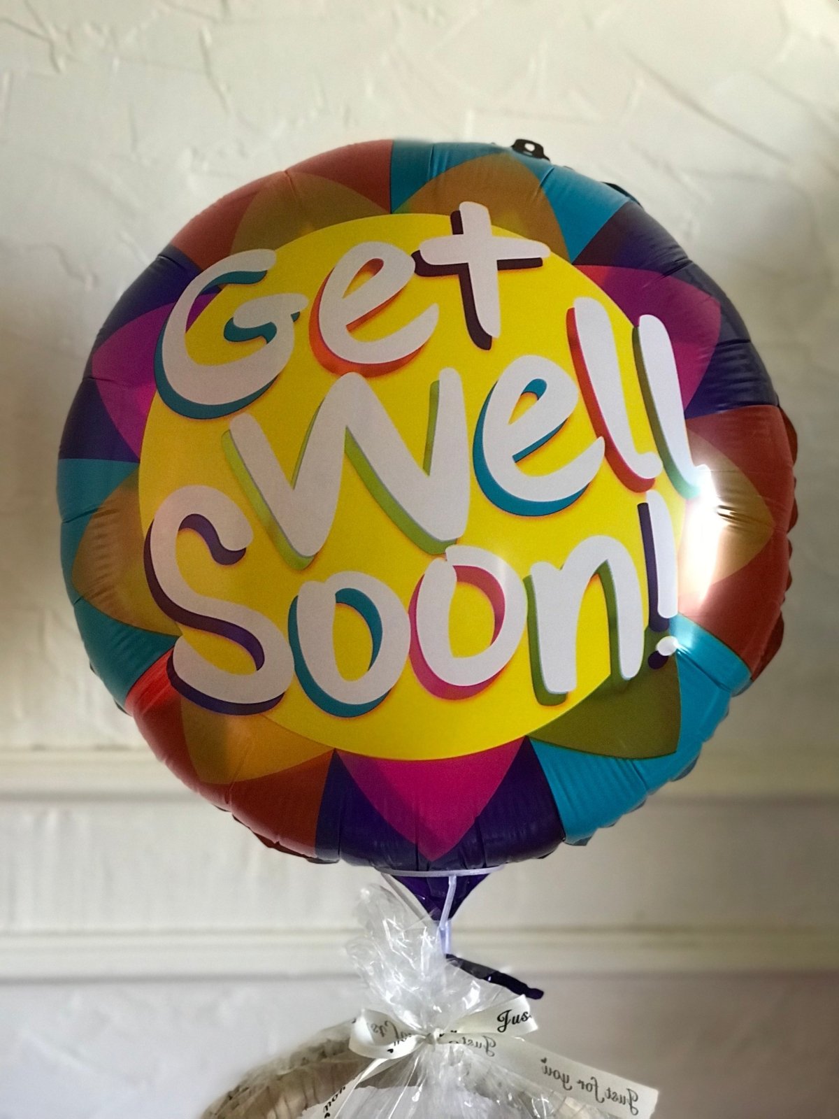 A cheerful Get Well Soon balloon with a smiling sun, rainbow rays, and multicolored text, tied to a wrapped gift, spreading positivity and warmth. Captured by our loyal customers.