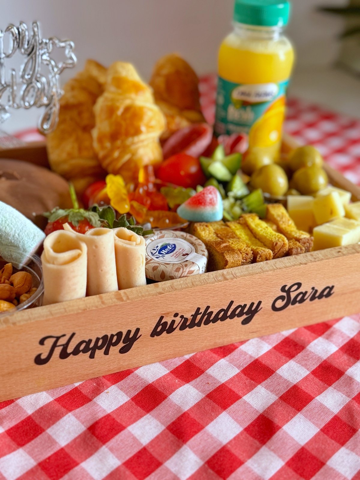 A vibrant assortment of pastries, fruits, nuts, and cheese on a personalized wooden tray with "Happy birthday Sara" text. Includes croissants, cheddar cheese, Turkish delights, candy, olives, and fresh juice for a cheerful breakfast experience.