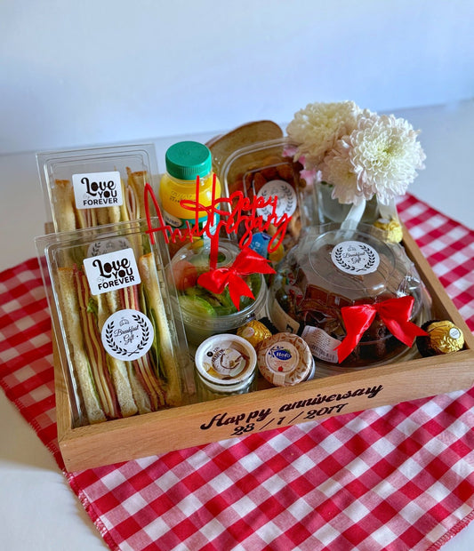 A beautifully arranged Happy Anniversary Breakfast tray featuring club sandwiches, volcano chocolate cake, assorted nuts, jam, butter, Fantancy chocolates, and a fresh juice bottle. The tray includes a personalized message with the date and a red "Happy Anniversary" topper, accented by a vase of white flowers and set on a red checkered cloth.