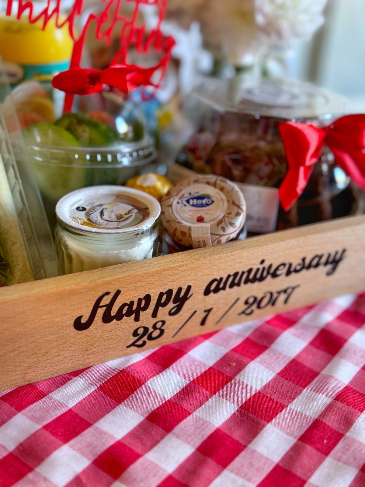 Close-up of the Happy Anniversary Breakfast tray displaying club sandwiches with "Love You Forever" stickers, cucumber, cherry tomatoes, volcano chocolate cake, and assorted nuts. The wooden tray is personalized with the anniversary date and decorated with a red "Happy Anniversary" topper and a vibrant red checkered tablecloth.