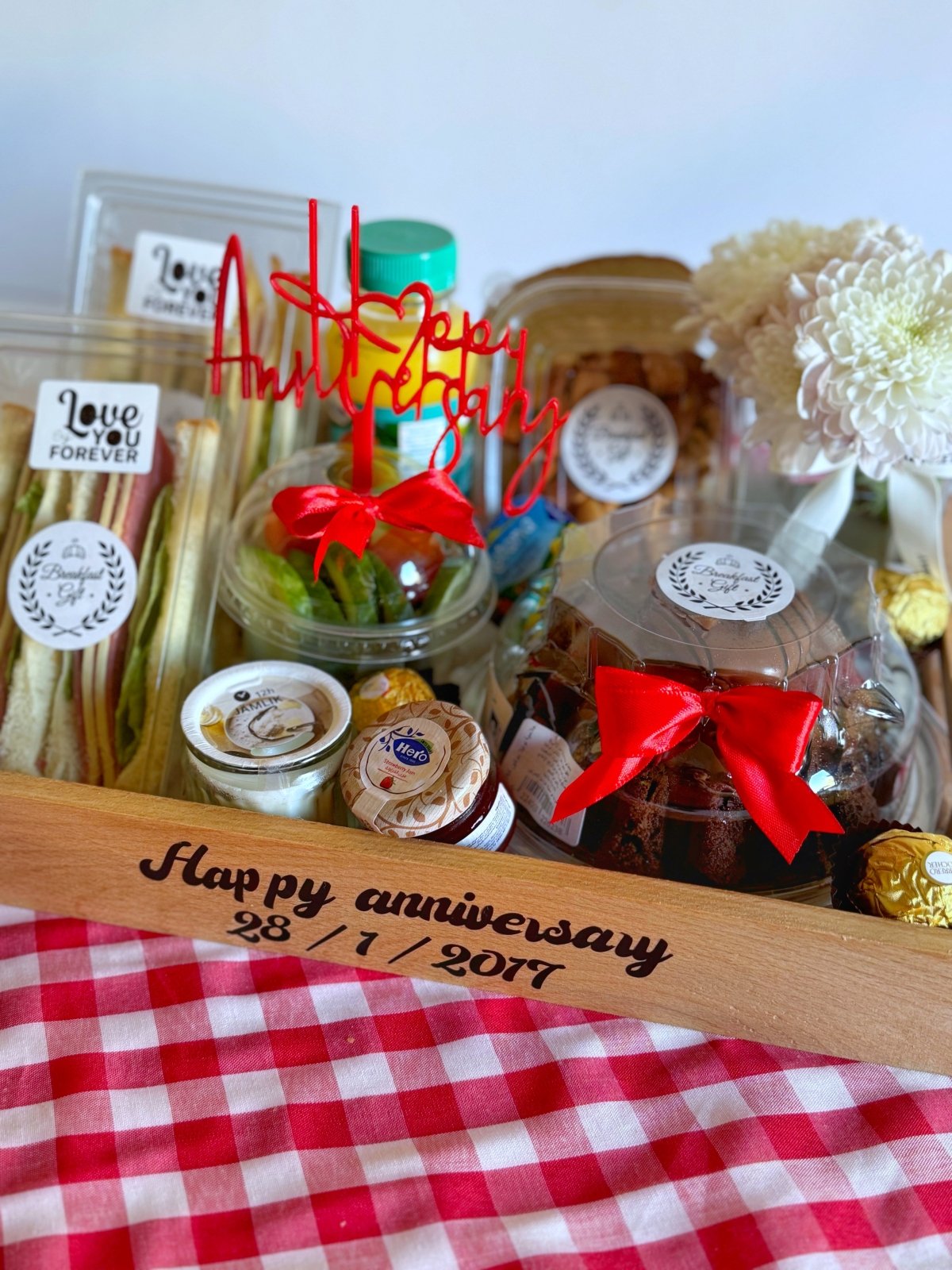 Complete view of the Happy Anniversary Breakfast tray with a decorative wooden base, featuring a variety of treats including volcano chocolate cake, club sandwiches, fresh juice, jam, butter, assorted nuts, and Fantancy chocolates. The display is enhanced with a red topper, white flowers in a vase, and a personalized anniversary message.