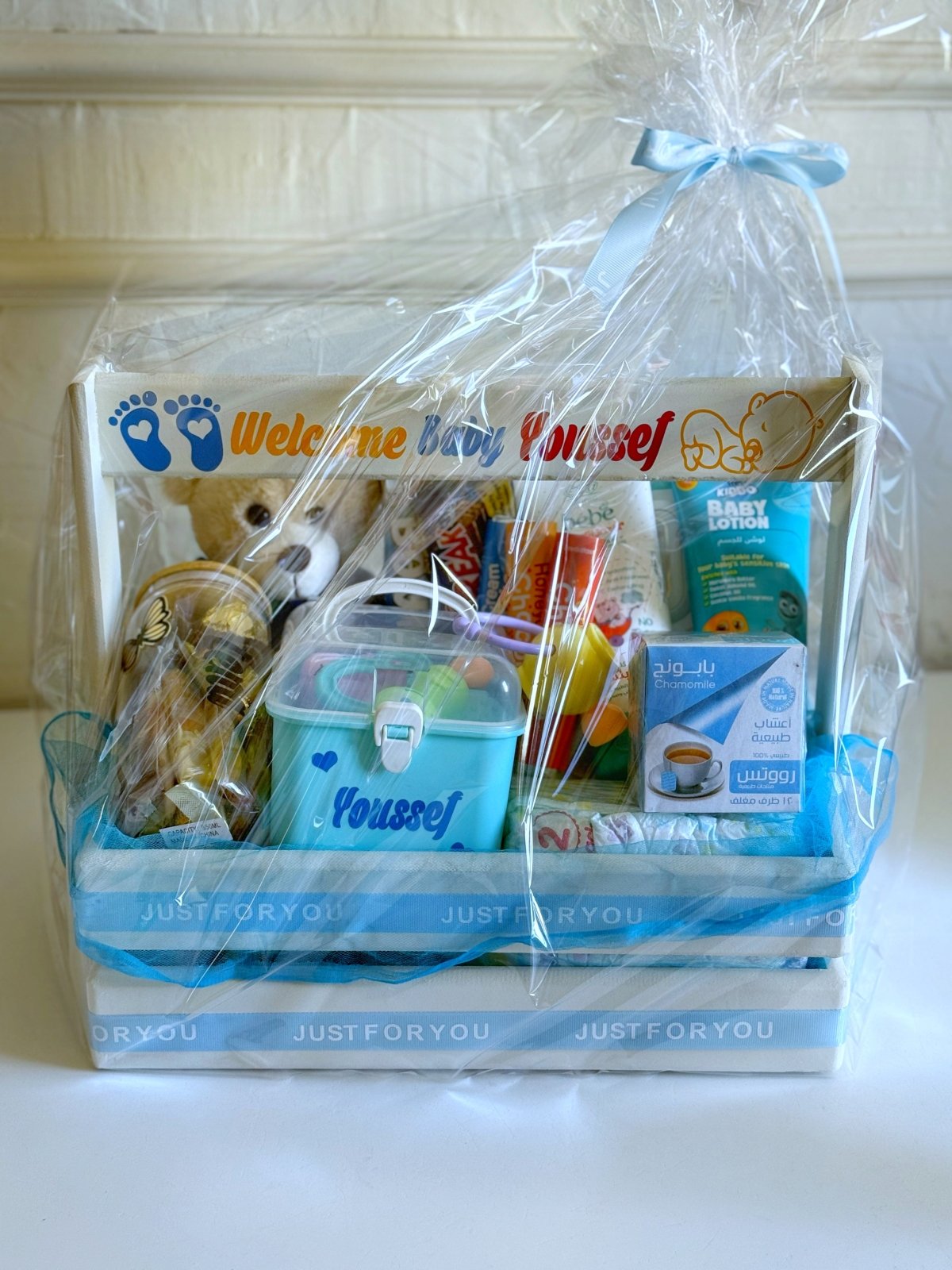 A wrapped baby boy gift basket in a white wooden crate with a blue ribbon and printed welcome message.