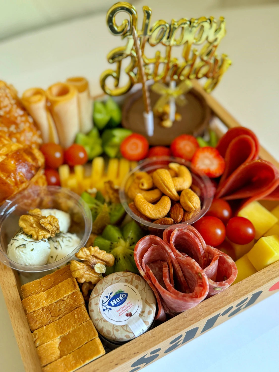 Close-up of salami, strawberries, kiwi slices, jam jar, and Happy Birthday topper in a wooden gift tray.