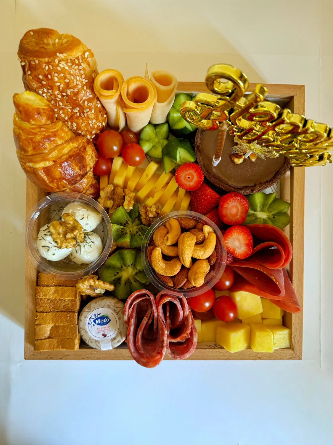 Top-down view of a personalized birthday breakfast tray with mini chocolate cake, fruits, turkey rolls, and mixed snacks.