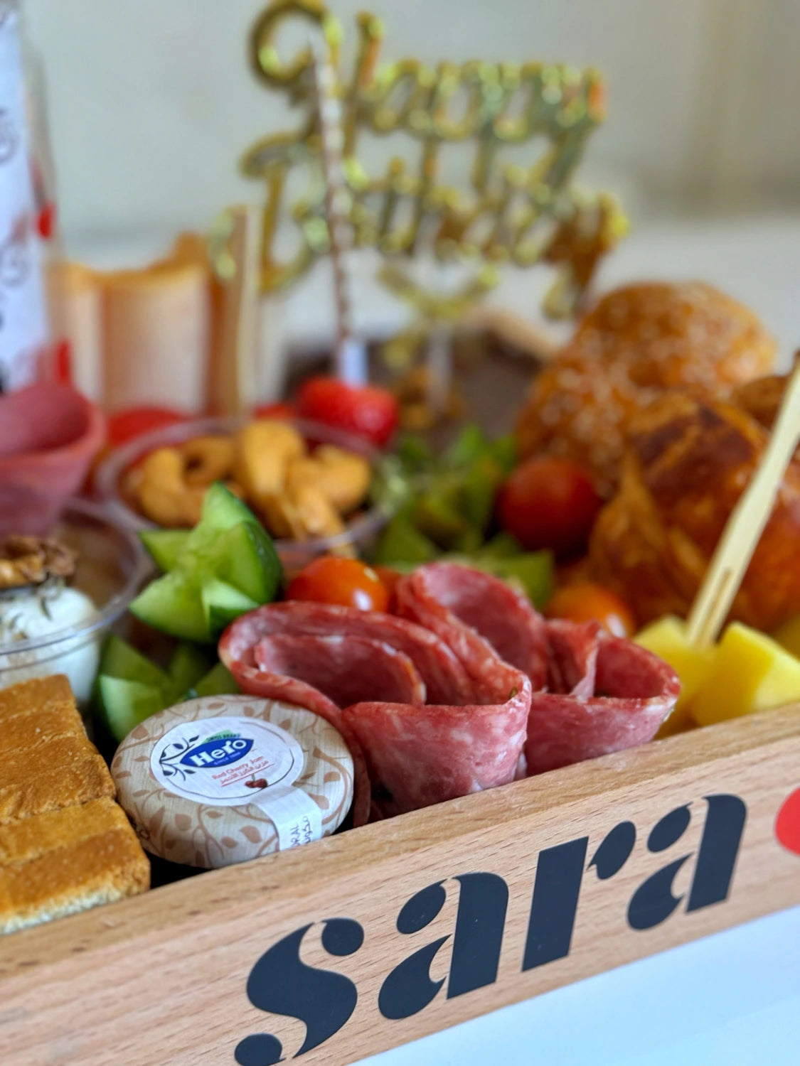 Close-up of salami, strawberries, kiwi slices, jam jar, and Happy Birthday topper in a wooden gift tray.