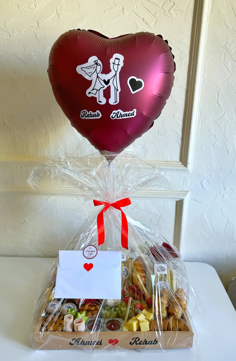 A heart-shaped balloon with bride and groom illustrations floats above a clear-wrapped breakfast tray labeled with the couple’s names.