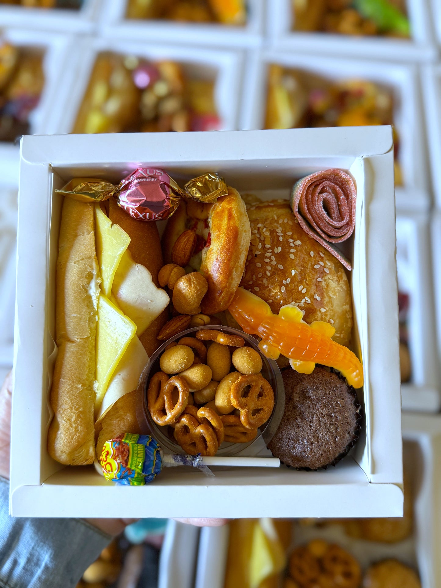 An open Mini Bites Birthday Box with a mini sandwich, paté, pizza, muffin, candy, pretzels, nuts, chocolate, and a lollipop with a name sticker.
