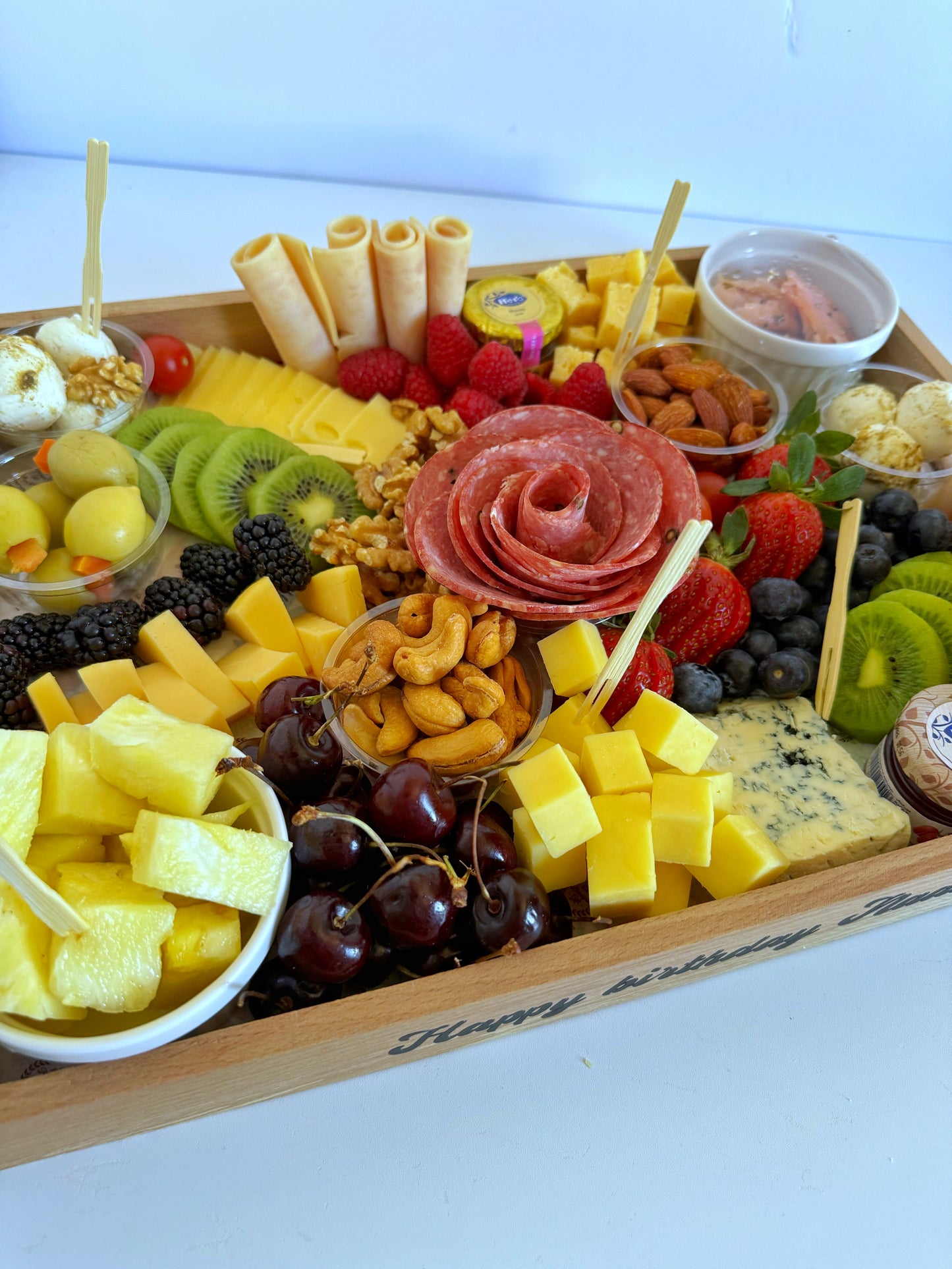 A luxurious wooden cheese board engraved with “Happy Birthday”, featuring gourmet cheeses, salami, fruits, and nuts, arranged in a celebratory layout.