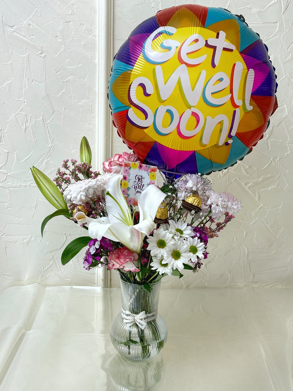 A cheerful glass vase with assorted fresh flowers, a colorful "Get Well Soon" balloon, Ferrero Rocher chocolates, and a floral greeting card—perfectly arranged to brighten someone's recovery.