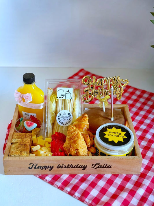 A beautifully arranged birthday breakfast tray with croissants, club sandwiches, fresh juice, zesty crumble canned cake, and a gold "Happy Birthday" topper, presented on a checkered tablecloth.