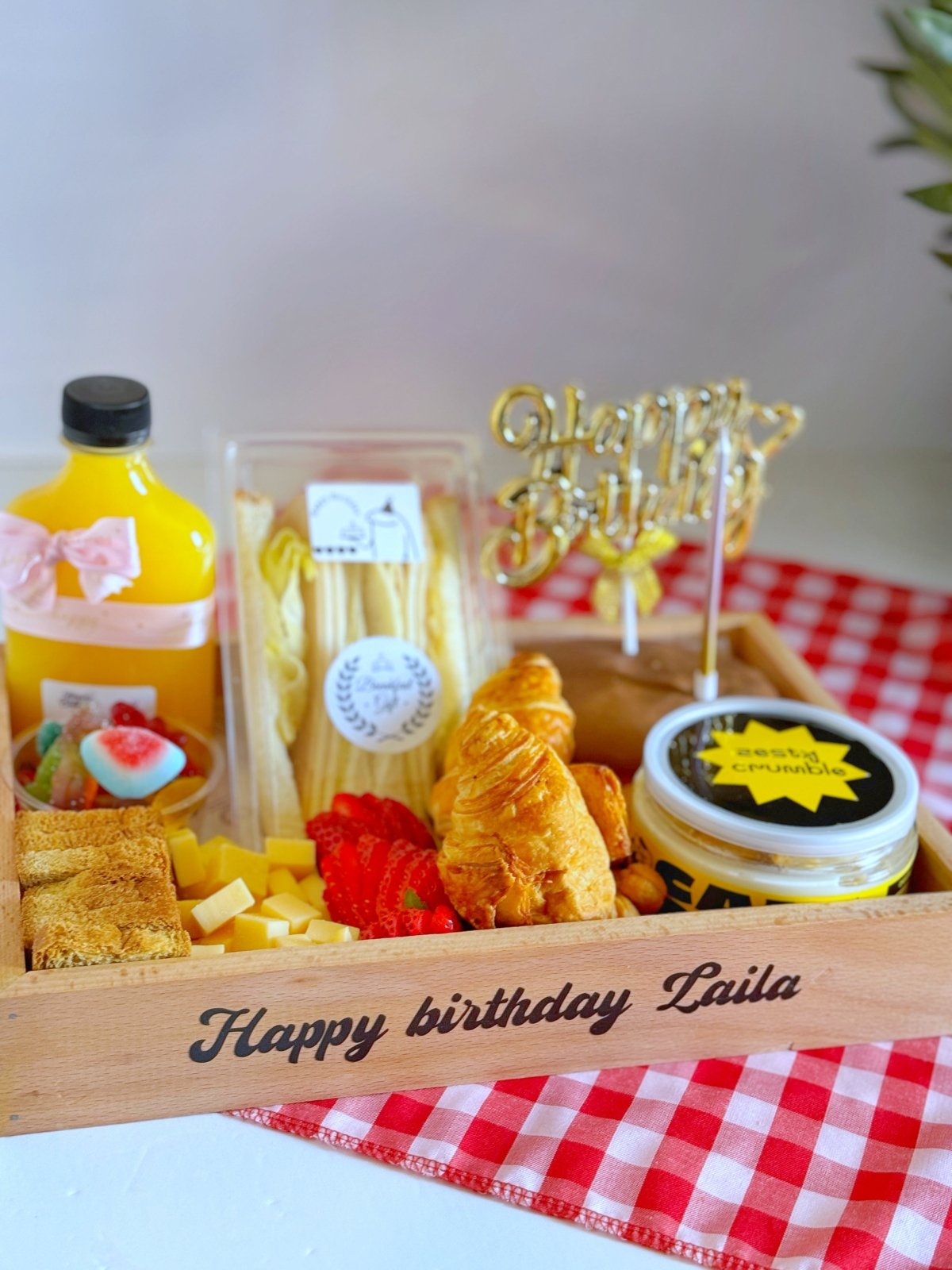 Close-up of a vibrant birthday breakfast tray featuring club sandwiches, croissants, cheese cubes, strawberries, zesty crumble canned cake, and a festive gold "Happy Birthday" topper.