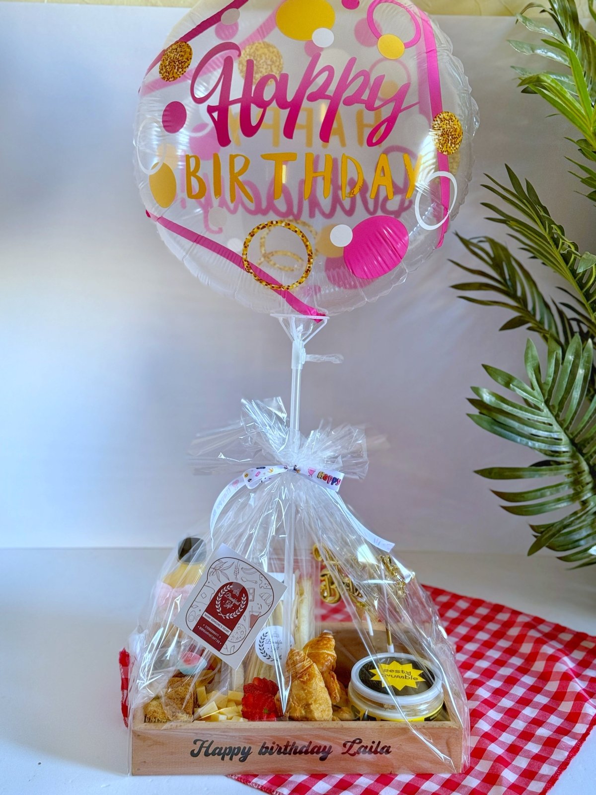 Trendy birthday breakfast tray wrapped elegantly with a transparent cover, topped with a pink and gold "Happy Birthday" balloon and a branded gift tag for a celebratory touch.