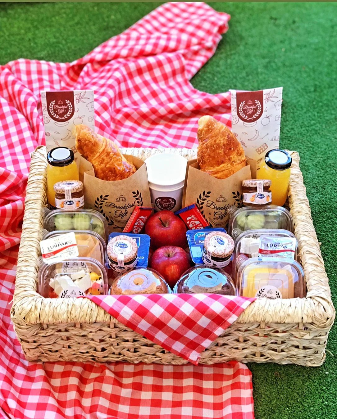Couples Basic Breakfast in a Basket for Two – a beautifully arranged rectangular wicker tray with croissants, toast, muffins, natural juices, fresh fruits, cheese, honey, jam, butter, Kit Kat chocolates, and tea. Thoughtfully presented with a checkered mat for a cozy and memorable breakfast experience.