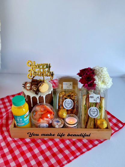 A beautifully presented Birthday Breakfast Cake & Candle gift set featuring a rich Nola chocolate cake with a golden 'Happy Birthday' topper, fresh fruit salad, assorted nuts, sandwiches, and a glowing candle in a wooden tray with a heartfelt message
