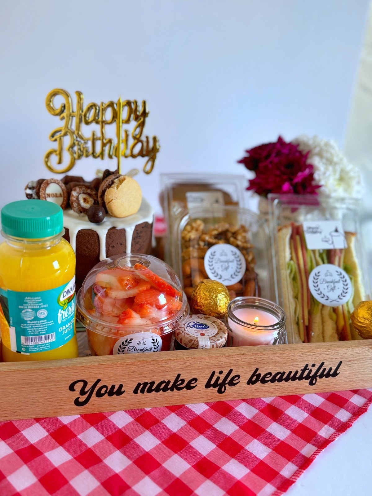 A delightful birthday breakfast tray with a chocolate cake, gourmet sandwiches, fresh juice, assorted nuts, and a glowing candle, styled on a red checkered cloth for a cozy and festive presentation.