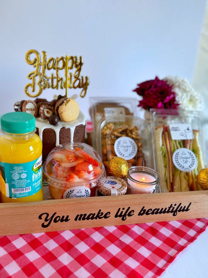 A delightful birthday breakfast tray with a chocolate cake, gourmet sandwiches, fresh juice, assorted nuts, and a glowing candle, styled on a red checkered cloth for a cozy and festive presentation.