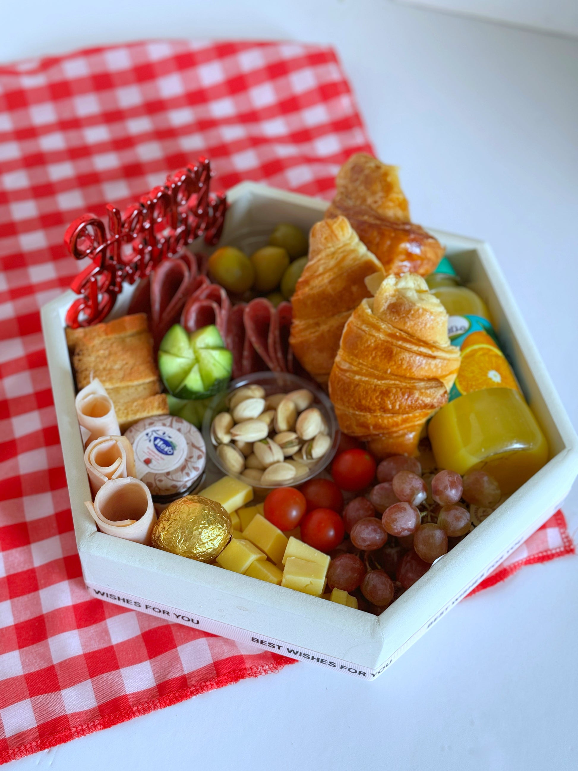 An exquisite birthday breakfast platter arranged in a unique hexagonal tray, featuring buttery croissants, fresh red grapes, cherry tomatoes, cheddar cheese cubes, cold cuts, stuffed olives, and gourmet extras like jam, butter, and Ferrero Rocher chocolates.