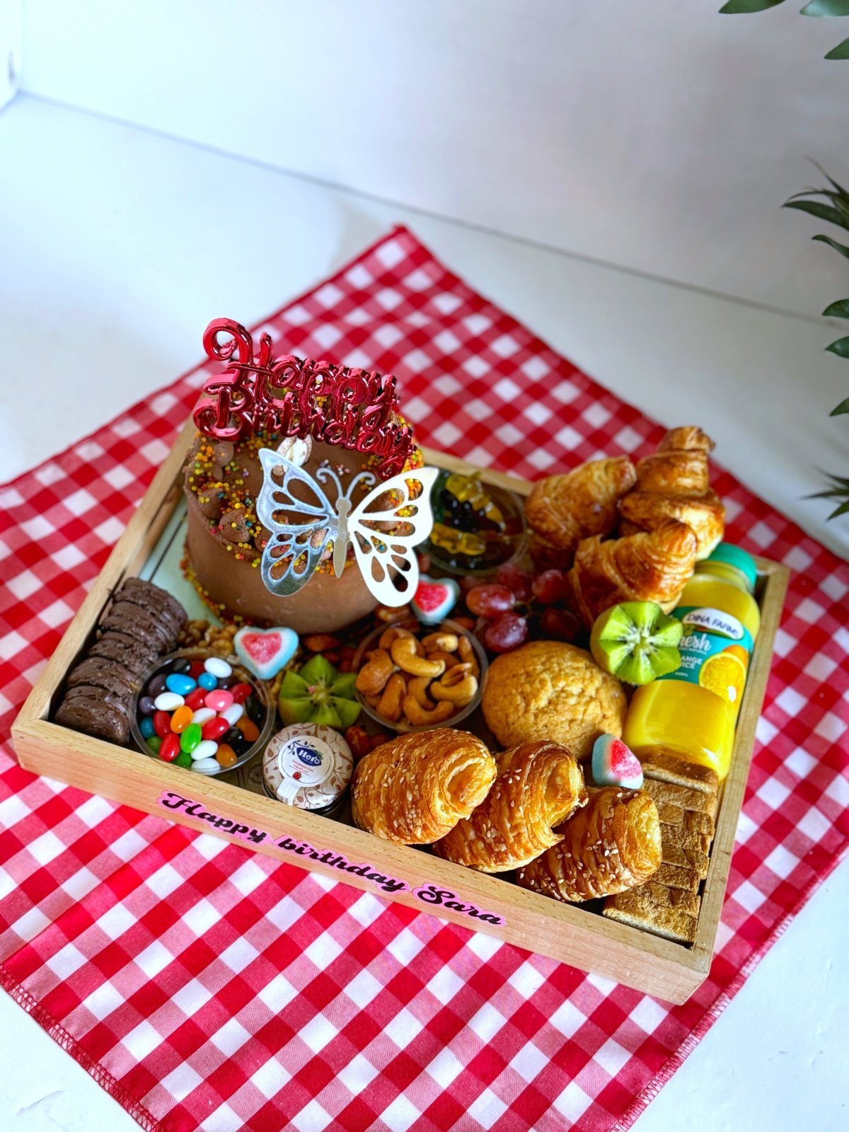 A vibrant Birthday Cake & Bakery gift set presented on a red checkered cloth, featuring a chocolate cake with festive sprinkles, fresh bakery treats, jellybeans, Hero jam, and Dina Farms juice for a joyful morning surprise.