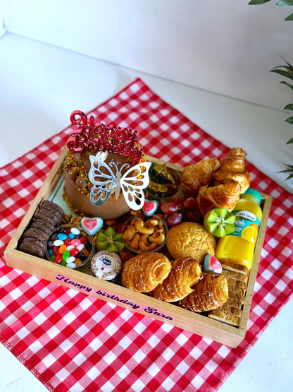 A vibrant Birthday Cake & Bakery gift set presented on a red checkered cloth, featuring a chocolate cake with festive sprinkles, fresh bakery treats, jellybeans, Hero jam, and Dina Farms juice for a joyful morning surprise.