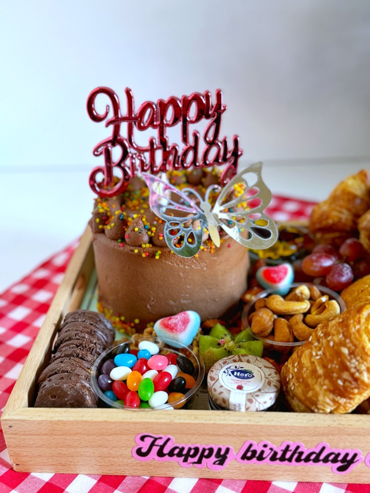 A luxurious wooden tray filled with a chocolate birthday cake, buttery croissants, sweet muffins, nuts, fresh fruits, and colorful candies, elegantly arranged with a 'Happy Birthday' topper for a memorable celebration.
