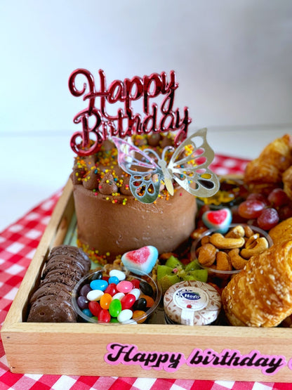 A luxurious wooden tray filled with a chocolate birthday cake, buttery croissants, sweet muffins, nuts, fresh fruits, and colorful candies, elegantly arranged with a 'Happy Birthday' topper for a memorable celebration.