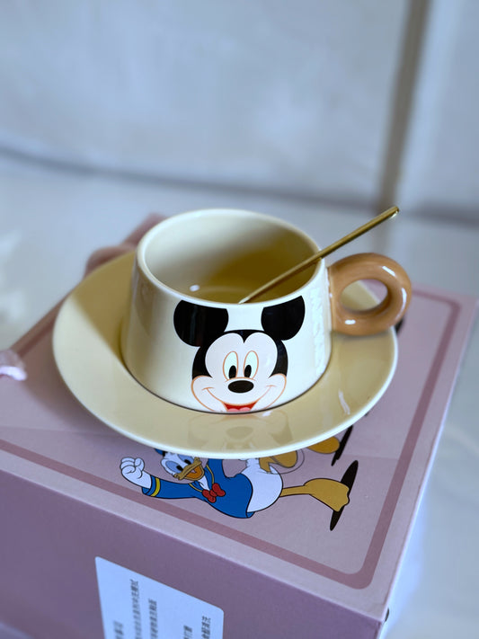 A cream-colored ceramic cup and saucer featuring Mickey Mouse, with a gold spoon. The gift box below shows Donald Duck’s artwork.