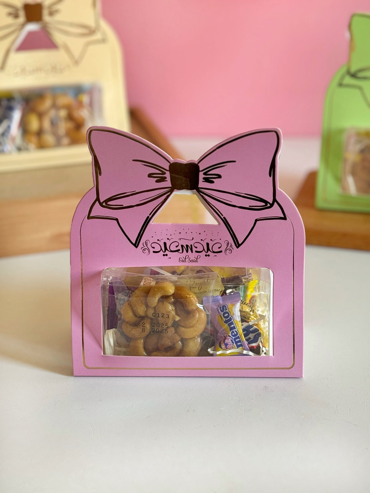 A pink Eid Bow Gift Box filled with chocolates, candies, and sugared cashews, featuring a clear display window and an elegant bow design.